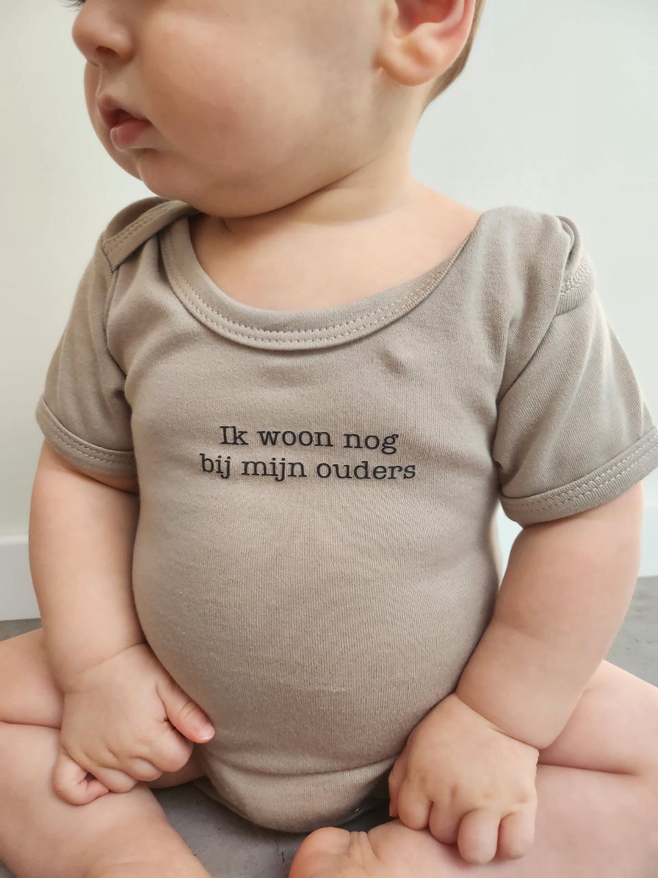 Baby met een bruine body waar de tekst 'ik woon nog bij mijn ouders' op staat. Geschikt als kraamcadeau, babyshower, verjaardagscadeau voor kind van 1 jaar